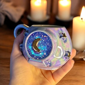 May include: A round, iridescent mug with a pearlescent finish featuring a central circular inlay filled with blue glitter and a gold crescent moon. The mug is decorated with small, raised gold star accents on its surface, creating a celestial theme.