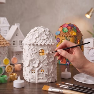 Könnte beinhalten: Eine weiße, hausförmige Keramiklaterne mit geprägten Blumenmustern, einer kleinen Bogentür und Fenstern. Das Objekt ist von kleinen Farbtöpfen, einer weißen Palette und mehreren Pinseln umgeben, wobei ein Licht aus dem Inneren leuchtet.