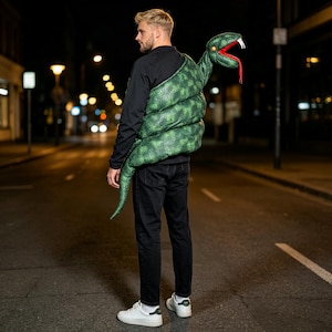 Könnte beinhalten: Ein grünes Schlangen-Accessoire aus Plüsch zum Tragen über der Schulter, mit texturiertem Schuppenmuster, einem roten offenen Maul mit weißen Fangzähnen und einem langen Schwanz. Die Schlange liegt auf dem Rücken einer schwarzen Jacke mit Jeans.