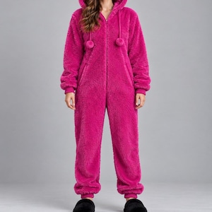 Può includere: Una tuta intera color fucsia brillante realizzata in materiale pile testurizzato e soffice. Il capo presenta una chiusura frontale con zip integrale, un cappuccio con due pon-pon pendenti su cordoncini e maniche lunghe con polsini.