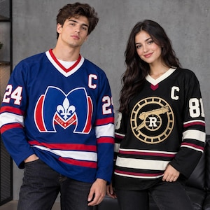 May include: A blue hockey jersey with a white and red M logo, the number 24, and a C patch, alongside a black hockey jersey featuring a gold cannon emblem, the number 81, and a C patch. Both jerseys have V-necks and striped sleeves.