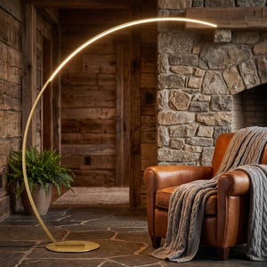 May include: A tall, arched floor lamp with a thin, gold-toned metal frame and a curved, integrated LED light strip that emits a warm glow. The lamp features a circular base and a sleek, minimalist design that curves over a leather armchair.