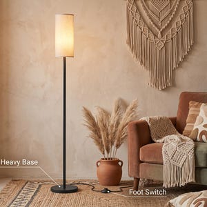 May include: A tall floor lamp with a slender black metal pole and a cylindrical off-white fabric shade. The lamp features a circular heavy base and a black cord with a foot switch. The text "Heavy Base" and "Foot Switch" are visible on the image.