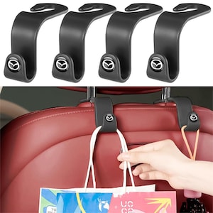 May include: Four matte black S-shaped car headrest hooks featuring a circular Mazda logo emblem on the lower curve. These hooks are designed to attach to the metal posts of a vehicle headrest to provide storage for bags and other items.