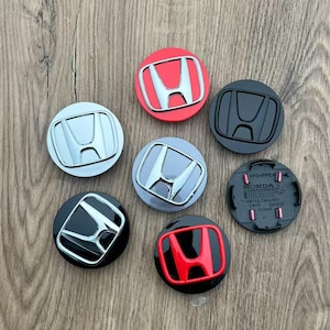 May include: A collection of seven circular Honda wheel center caps in various finishes, including chrome, matte black, red, and silver. One cap is flipped over to reveal the black plastic underside with molded text and pink clips.