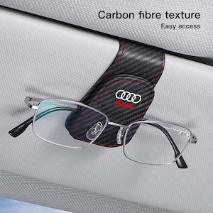 Peut inclure: Un porte-lunettes de pare-soleil de voiture noir avec une texture fibre de carbone et des coutures rouges. Le support présente le logo Audi blanc et le texte Audi rouge. Des lunettes à monture argentée sont fixées. Texte : Carbon fibre texture.