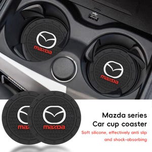 May include: Two circular black silicone car cup coasters featuring the white Mazda logo and red Mazda text. The coasters have a textured honeycomb pattern and a notched outer rim. The text Mazda series Car cup coaster is visible.