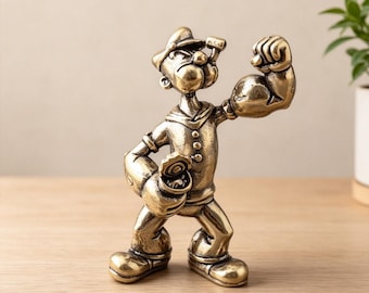 Op de afbeelding: Een metalen, bronskleurig beeldje van een zeeman met een pet en uniform. De figuur heeft één arm gebogen om een grote biceps te tonen en houdt een open blikje in de andere hand. Het personage heeft een pijp in de mond en een vastberaden uitdrukking.