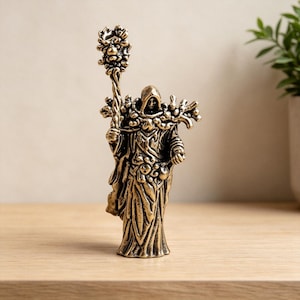 Puede incluir: Una figura metálica de color bronce que representa a una persona encapuchada con una túnica larga y fluida. La figura sostiene un bastón alto y ornamentado rematado con un grupo de esferas y presenta detalles intrincados en la superficie.