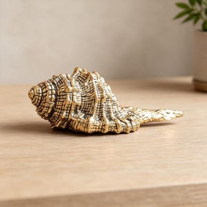 Puede incluir: Una figura de concha marina en tono dorado metálico con una superficie texturizada muy detallada, que presenta crestas intrincadas y un patrón en espiral. El objeto tiene un acabado pulido que refleja la luz, mostrando una forma de caracola realista.