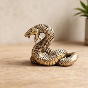 Puede incluir: Una figura de serpiente de metal en tono dorado con detalles intrincados de escamas. La serpiente se muestra en una posición enroscada con la cabeza levantada y la boca ligeramente abierta, exhibiendo una superficie texturizada y un acabado pulido.