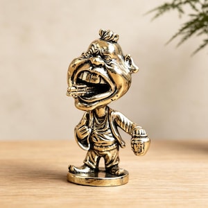Könnte beinhalten: Eine messingfarbene Metallfigur eines cartoonartigen Charakters mit überdimensionalem Kopf, weit geöffnetem Mund und einer Zigarre. Die Figur trägt eine Jacke und eine Hose und hält einen kleinen Krug mit dem chinesischen Schriftzeichen 酒 in der Hand.