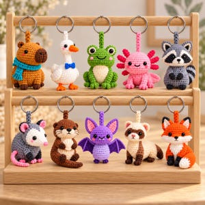 May include: A collection of ten crocheted animal keychains displayed on a two-tiered wooden stand. The figures include a capybara, goose, frog, axolotl, raccoon, opossum, otter, bat, ferret, and fox, each attached to a silver metal ring.