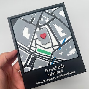 Puede incluir: Un marco negro cuadrado que contiene un mapa en 3D con edificios grises, calles blancas y una zona de parque verde. Un pequeño corazón rojo está colocado sobre un edificio central. El borde negro inferior presenta el texto blanco: Fran&Paula 04/07/2026 40 94582020907901, -4 120679209640419.