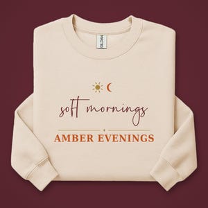 Peut inclure: Un sweat-shirt à col rond beige avec l'inscription "soft mornings" en police cursive rouge foncé au-dessus du texte "AMBER EVENINGS" en lettres capitales orange. Une petite icône de soleil et de croissant de lune est centrée au-dessus.
