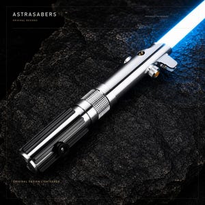 Puede incluir: Una empuñadura de sable de luz de metal plateado con detalles de agarre verticales en negro y una hoja azul brillante. La empuñadura presenta una sección central texturizada y moleteada, un botón circular dorado y un pequeño interruptor negro.