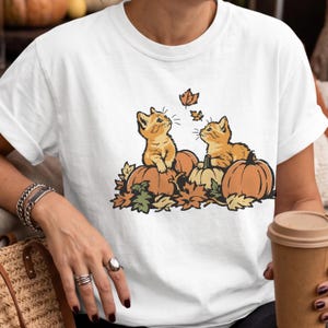 Op de afbeelding: Een wit t-shirt met een grafische illustratie van twee oranje kittens die tussen pompoenen en herfstbladeren zitten. Eén kitten kijkt naar een vallend blaadje. Het ontwerp gebruikt warme tinten oranje, bruin en groen voor het tafereel.