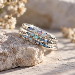 Op de afbeelding: Een stapel van drie zilveren ringen, elk voorzien van een kanaal met kleine, ruwe, iriserende opaalstenen die flitsen van blauw, groen en roze vertonen. De metalen banden hebben een getextureerde, organische afwerking met een metaalglans.