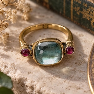 May include: A gold ring featuring a large, cushion-cut, translucent blue-green gemstone at the center, flanked by two smaller, round, faceted deep red gemstones. The metal band has a textured finish and a decorative border around the central stone.