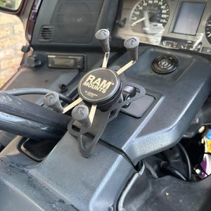 Pode incluir: Um suporte de telefone RAM Mounts X-Grip preto montado no painel de uma motocicleta. O dispositivo possui um cubo central circular com o texto RAM MOUNTS X-GRIP e quatro braços ajustáveis com pontas de borracha, cada um marcado com um logotipo R.