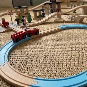 May include: A wooden toy train set featuring a red two-car train on a circular track with light blue and natural wood segments. The set includes a station, a bridge, green trees, and small figures positioned near a railroad crossing sign.