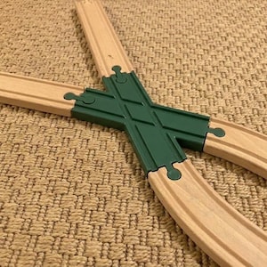 May include: A wooden toy train track intersection piece featuring a central dark green plastic X-shaped crossing connector. The connector joins four light-colored wooden track segments, each with a grooved center and interlocking puzzle-style ends.