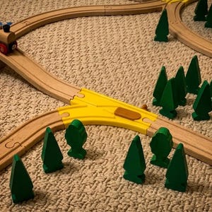 Könnte beinhalten: Ein hölzernes Spielzeugeisenbahn-Set mit hellbraunen Schienen, einer gelben Kunststoff-Weiche, einer roten Brücke und einer kleinen Lokomotive. Das Set enthält mehrere grüne Holzbäume in verschiedenen Formen, die um die Gleise angeordnet sind.
