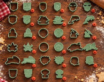 May include: A collection of sage green plastic cookie cutters in various shapes, including animals, shields, and circular emblems, arranged in a grid on a wooden surface with small orange circular markers placed next to several of the cutters.