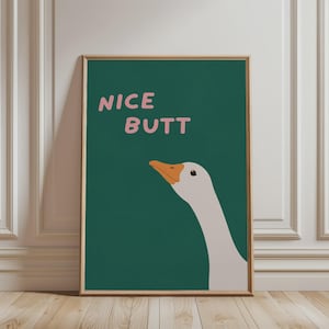 Op de afbeelding: Een rechthoekige wandkunstprint in een lichthouten lijst met een donkergroene achtergrond en de kop en nek van een witte gans. De tekst NICE BUTT staat in lichtroze, afgeronde, schreefloze hoofdletters boven de illustratie van de gans.