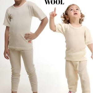 May include: Two sets of cream-coloured short-sleeved shirts and long trousers made of wool. The garments feature a ribbed texture, a round neckline, and a simple design. The word WOOL is printed in black text above the right-side set of clothing.