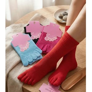 May include: A pair of solid red toe socks worn on feet, with additional pairs of toe socks in light blue, magenta, and black packaged with pink cardboard headers. Each pair features individual compartments for every toe and a ribbed knit texture.