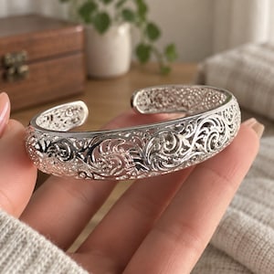 May include: A silver-toned cuff bracelet featuring an intricate, openwork filigree pattern of swirling, interconnected scroll designs. The metal band has a polished, reflective finish and a wide, C-shaped opening for easy wear on the wrist.