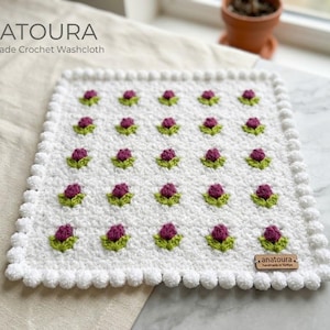Puede incluir: Un paño de cocina rectangular de ganchillo blanco con un patrón repetitivo de pequeñas flores moradas y hojas verdes. El borde consiste en pompones de ganchillo blancos. Una etiqueta de madera tiene grabado el texto anatoura handmade in Türkiye.