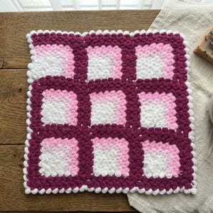 Puede incluir: Un paño de cocina cuadrado de ganchillo con un patrón de cuadrícula de nueve cuadrados. Cada cuadrado muestra un efecto degradado que va del blanco al rosa claro y al magenta oscuro. El borde tiene un acabado de punto bobble blanco.