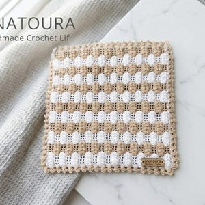 Puede incluir: Un paño de ganchillo cuadrado con rayas horizontales alternas de puntos de burbuja texturizados en beige y blanco. Los bordes están rematados con un borde decorativo beige y una pequeña etiqueta marrón con la inscripción ANATOURA.