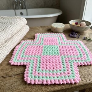 Puede incluir: Una manopla de baño de ganchillo cuadrada con un patrón en forma de cruz en hilo rosa pastel y verde menta alternados. El punto texturizado crea una superficie en relieve y los bordes están rematados con un borde festoneado en hilo rosa.
