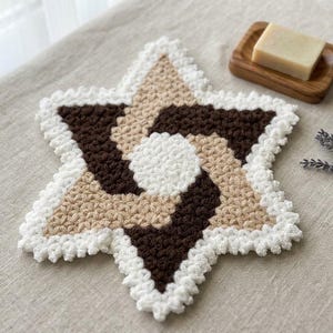 Puede incluir: Un posavasos o paño de crochet en forma de estrella con un patrón de remolinos en hilo marrón oscuro, beige y blanco. El centro es blanco, rodeado por secciones alternas de marrón oscuro y beige, acabado con un borde festoneado blanco.
