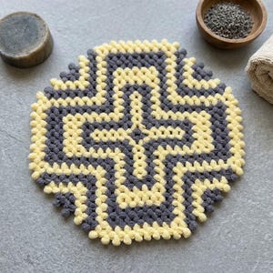 Puede incluir: Una alfombrilla de ganchillo circular con un patrón texturizado de puntos de burbuja en anillos concéntricos alternos de hilo amarillo pálido y gris oscuro. El diseño crea un motivo geométrico en forma de cruz que irradia hacia el borde.