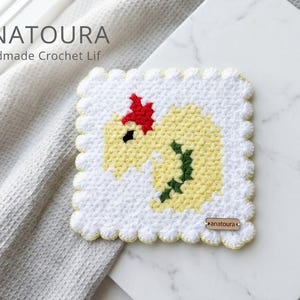 Puede incluir: Un posavasos cuadrado de ganchillo con un borde festoneado blanco y un diseño central de un dragón amarillo con cresta roja y espina dorsal verde oscuro. El posavasos tiene una pequeña etiqueta de madera con la marca "anatoura" abajo.