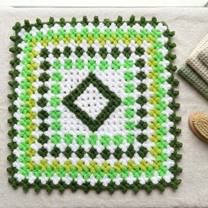 May include: A square crochet washcloth featuring a geometric pattern in white, lime green, and dark forest green yarn. The design consists of concentric diamond shapes with a textured, puffy stitch finish and a decorative scalloped border edge.