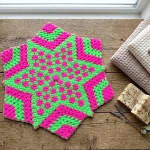 Puede incluir: Un agarrador de cocina hexagonal tejido a ganchillo con un vibrante patrón de estrella en hilo rosa neón y verde lima brillante. El diseño geométrico texturizado está creado con puntos densos y elevados, formando un motivo de estrella audaz.