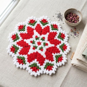 Puede incluir: Un tapete de ganchillo redondo con un patrón en forma de estrella en hilo rojo vibrante, blanco y verde bosque. El diseño intrincado consiste en puntos agrupados que forman un motivo floral con un borde festoneado y una pequeña etiqueta de madera.