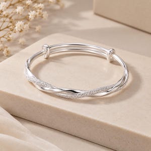 May include: A silver-toned adjustable bangle bracelet featuring a twisted design. One strand has a smooth, polished metallic finish, while the other strand has a textured, sparkling surface. The bracelet includes two small sliding loops for sizing.