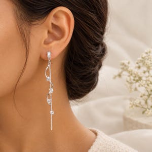 Op de afbeelding: Een lange zilverkleurige oorhanger met een fijne, dunne ketting en een parallelle golvende metalen draad. Het ontwerp is versierd met verschillende kleine, heldere en sprankelende zirkonia-steentjes die langs de oorhanger zijn geplaatst.