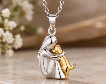 Könnte beinhalten: Eine silberfarbene Anhängerkette mit einer stilisierten Figur, die einen kleinen goldfarbenen Hund umarmt. Der Anhänger hängt an einer zarten silbernen Kette und zeigt eine glatte, polierte Oberfläche sowohl an der menschlichen Figur als auch am Hund.