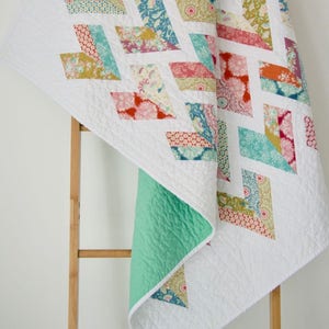 May include: A white quilt with a chevron pattern in shades of pink, orange, teal, and yellow. The quilt is folded and draped over a wooden ladder.