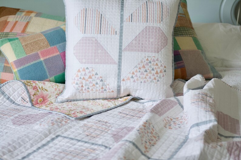 Pu&ograve; includere: Un copriletto bianco trapuntato con un motivo geometrico in tonalit&agrave; rosa e grigio. Il copriletto &egrave; ripiegato per rivelare un tessuto con motivo floreale sottostante. Due cuscini con motivi geometrici sono sul letto.