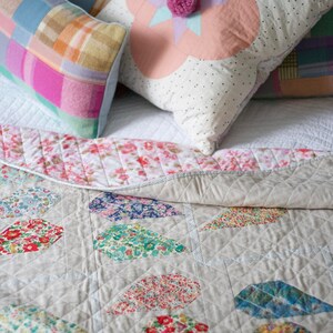 Pu&ograve; includere: Un trapunta di patchwork bianco e grigio con un motivo floreale. La trapunta &egrave; piegata e ha un cuscino bianco con pois rosa e bianchi e un fiore rosa con una stella al centro. Ci sono altri due cuscini con disegni di patchwork rosa, blu e verde.