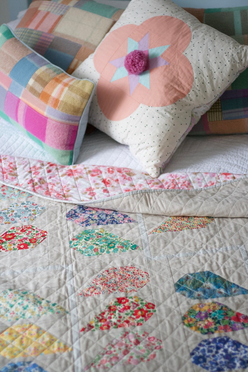 Pu&ograve; includere: Un trapunta di patchwork bianco e grigio con un motivo floreale. La trapunta &egrave; piegata e ha un cuscino bianco con pois rosa e bianchi e un fiore rosa con una stella al centro. Ci sono altri due cuscini con disegni di patchwork rosa, blu e verde.