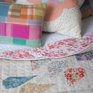 Pu&ograve; includere: Un piumino bianco con un design a patchwork che presenta motivi floreali in tonalit&agrave; di rosa, blu e verde. Il piumino &egrave; ripiegato su un letto con diversi cuscini, tra cui uno con un motivo a pois rosa e bianco e un altro con un motivo a quadri in tonalit&agrave; di rosa, blu e marrone.